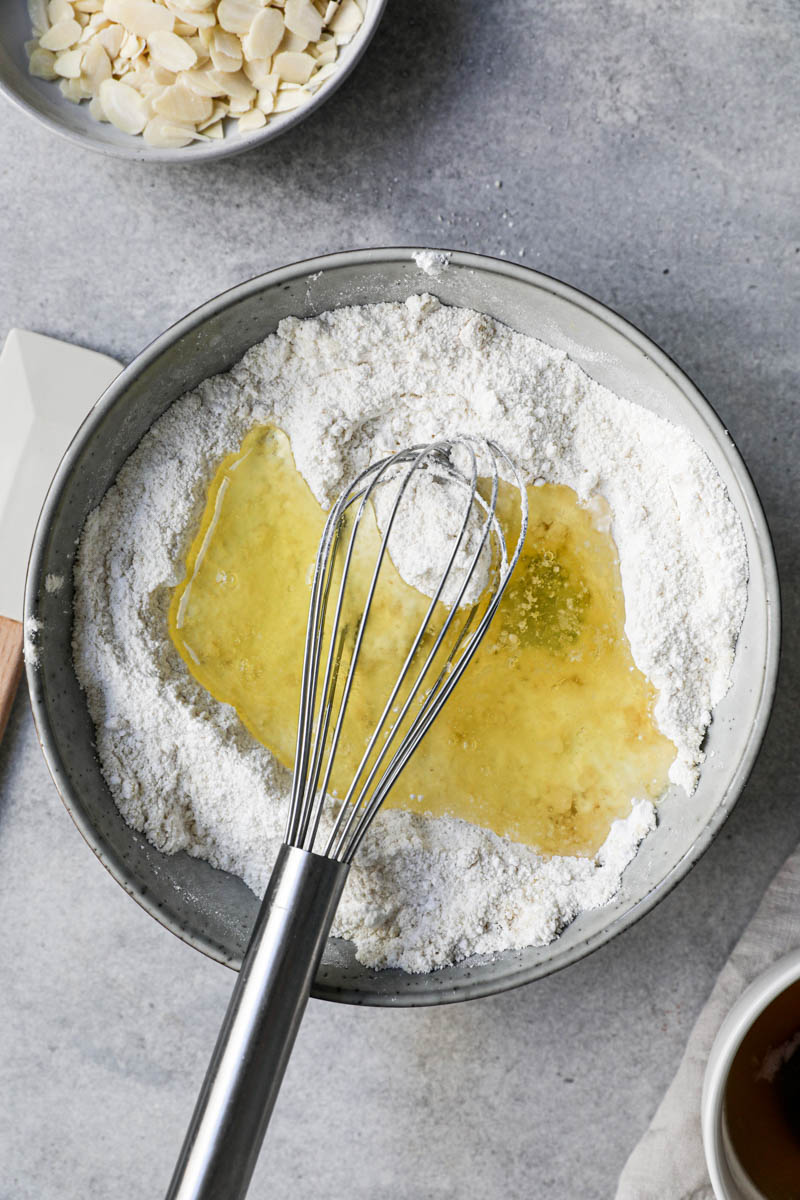 Los ingredientes secos mezclados dentro de un bol gris con un batidor dentro, un plato lleno de almendras arriba del cuadro y claras de huevo dentro de un bol a su lado.