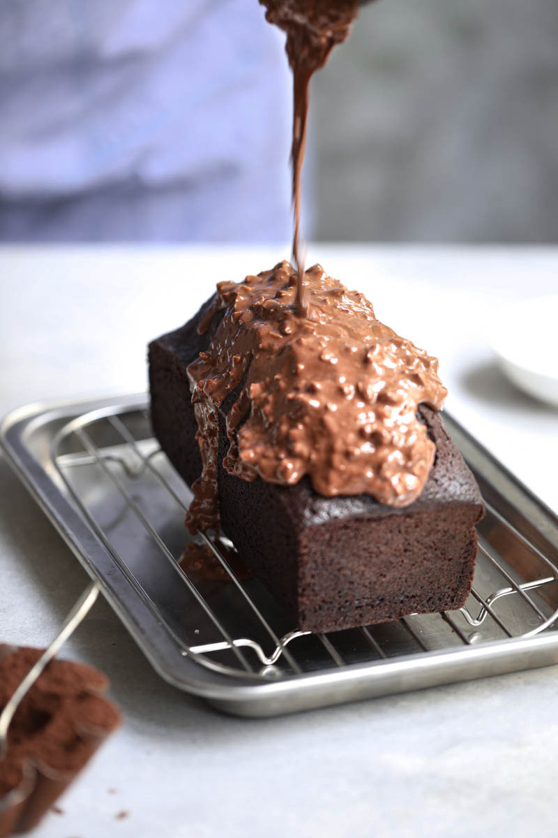 El bizcocho de chocolate horneado sobre una rejilla y una mano que está cubriendo el glaseado con una jarrita gris.