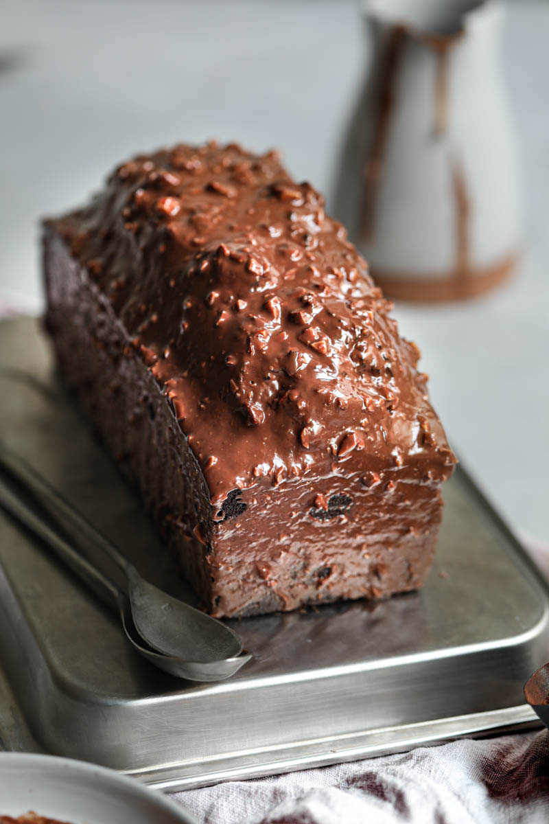 El budín de chocolate glaseado sobre una bandeja para horno plateada invertida con una jarrita por detrás.
