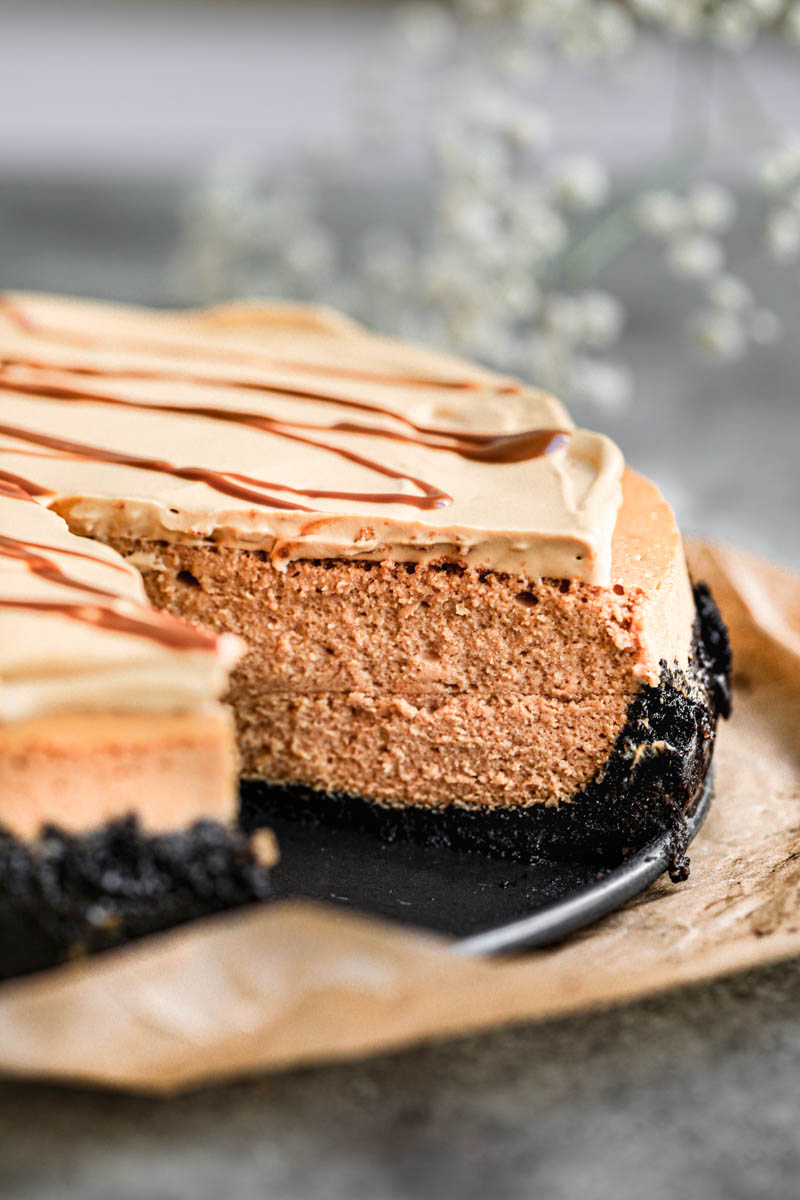 Primer plano del cheesecake de dulce de leche cortada viendo el interior sobre un plato negro colocado sobre un pedazo de papel vegetal marrón con algunas flores borrosas por detrás.