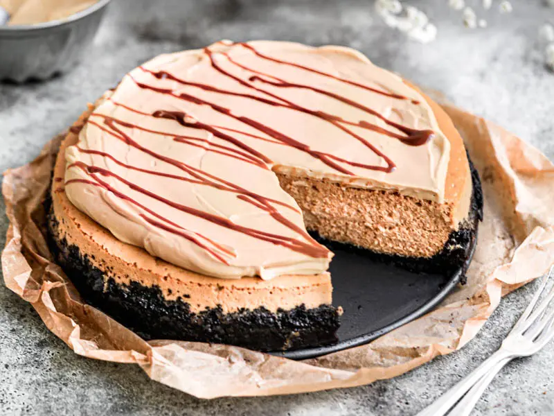 Primer plano del cheesecake de dulce de leche cortada viendo el interior sobre un plato negro colocado sobre un pedazo de papel vegetal marrón con algunas flores borrosas por detrás.