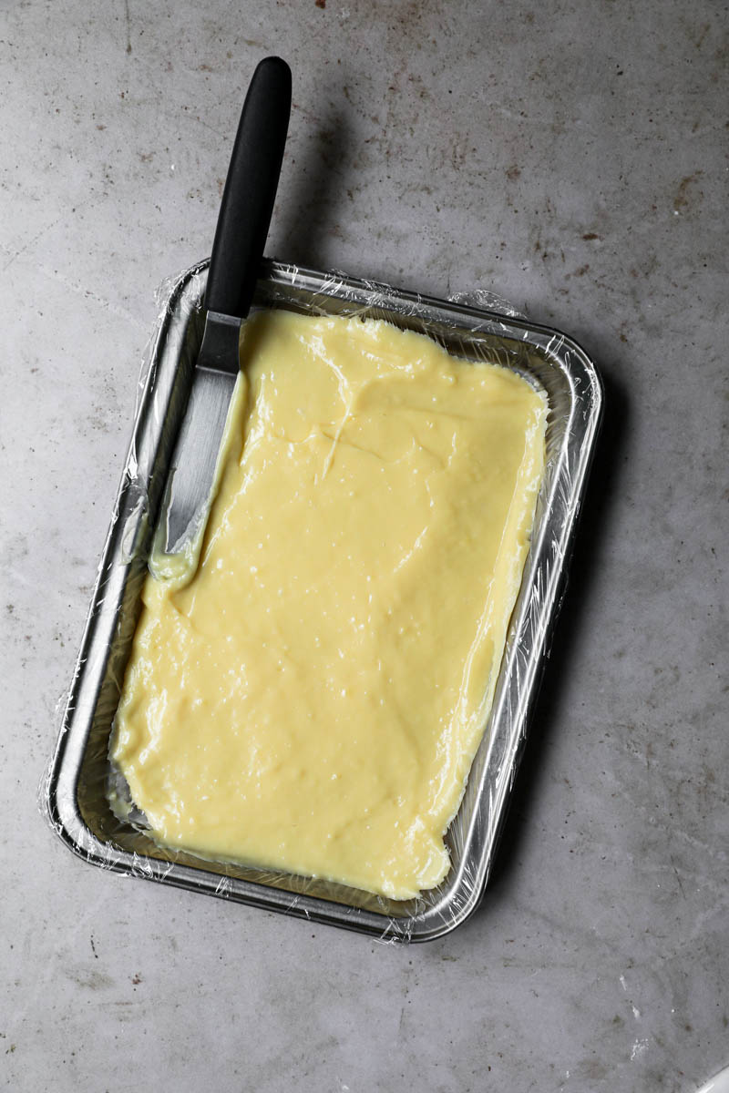 La crema pastelera en una pequeña bandeja forrada con papel film y una espátula pequeña en un ángulo.