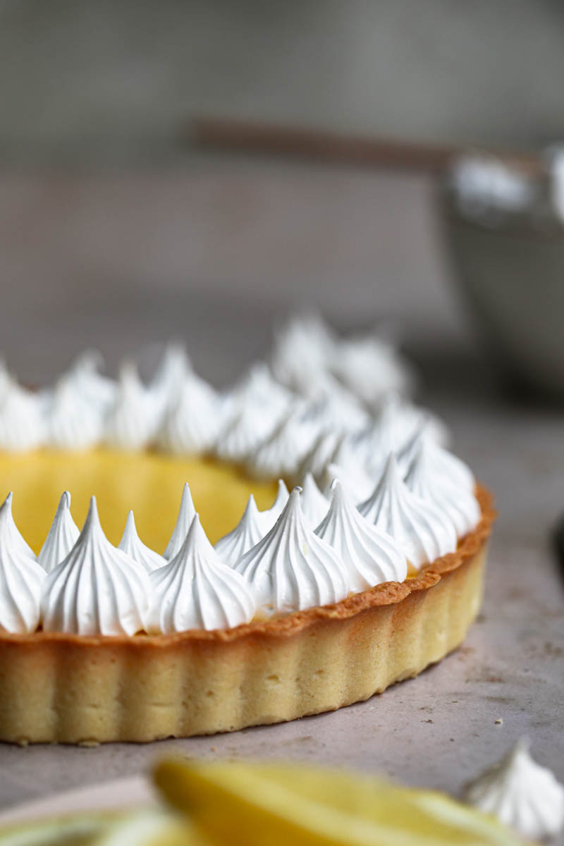 Mitad de la tarta de limón cubierta con merengue suizo con un bol pequeño y una espátula por detrás.