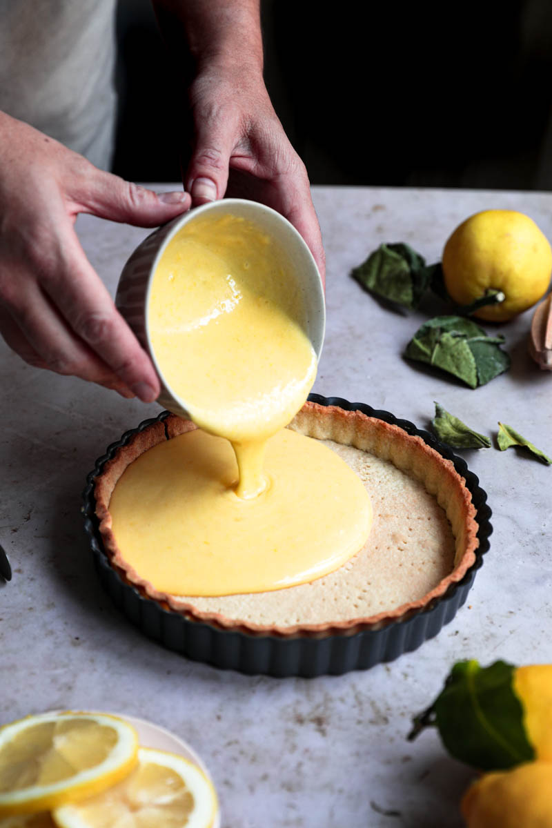 Dos manos vertiendo el curd de limón listo sobre la masa de la tarta que está rodeada de limones.