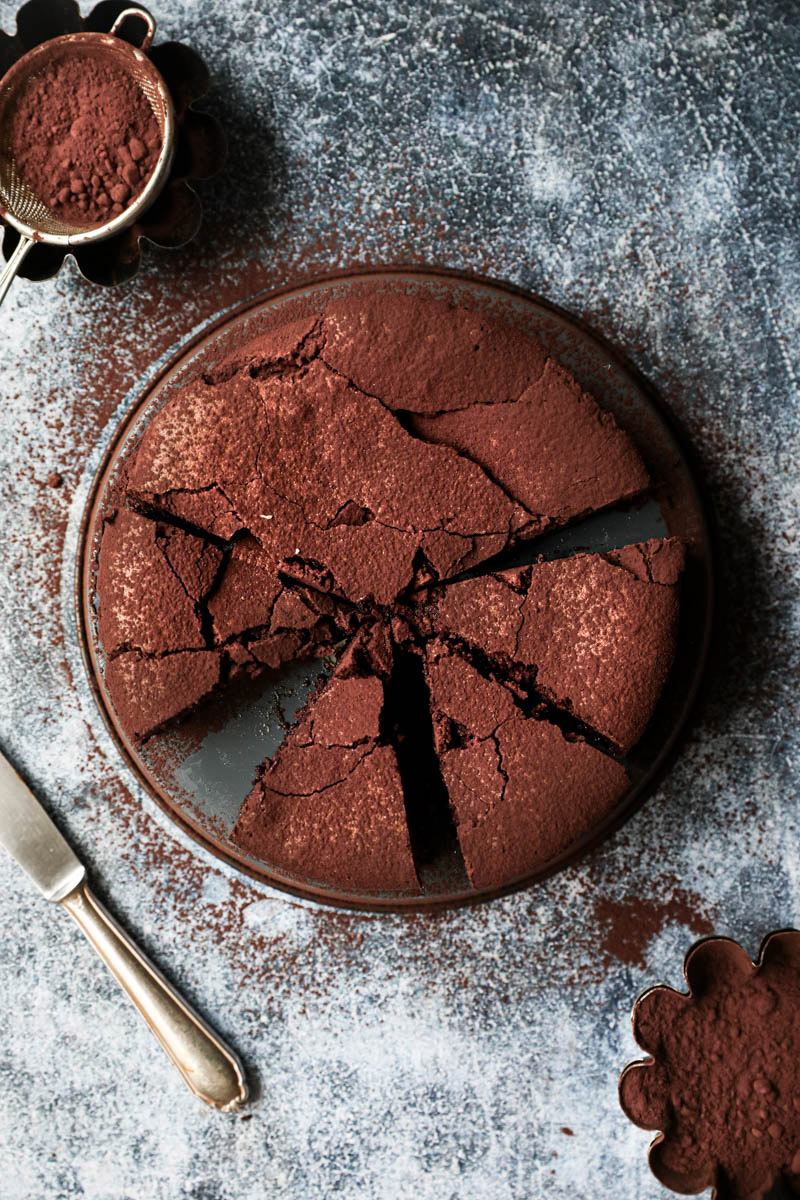 La torta húmeda de chocolate cortada sobre una bandeja gris invertida con un cuchillo al costado y bol con cacao al costado visto desde arriba.