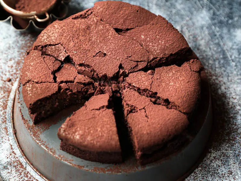 La torta húmeda de chocolate cortada sobre una bandeja gris invertida con un cuchillo al costado y bol con cacao detrás.