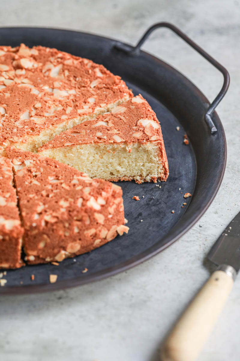 The sliced almond cake on a black tray with a knife on the side as seen from the side.