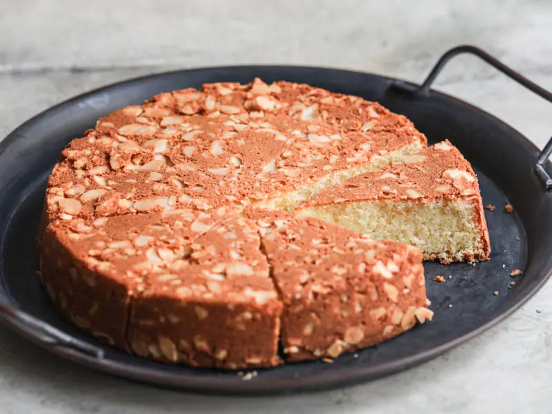 The sliced almond cake on a black tray with a knife on the side as seen from the side.