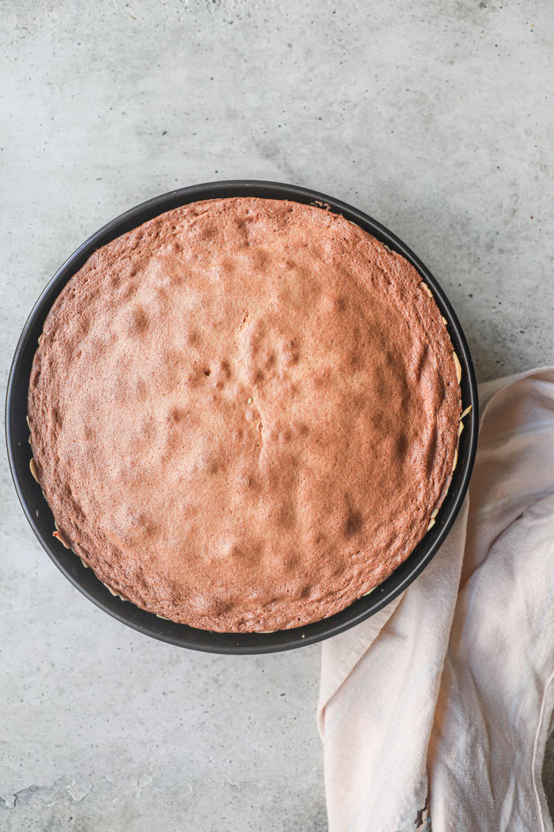 The baked almond paste cake inside the cake tin with a kithen towel on the side.