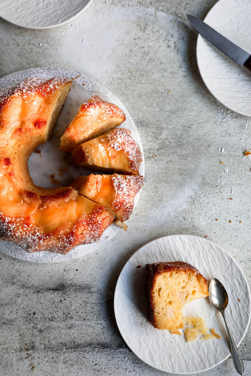 Overhead shot of the sliced apple cake on a marble stand surrounded by plates and a slice of cake on a white plate in the bottom right corner