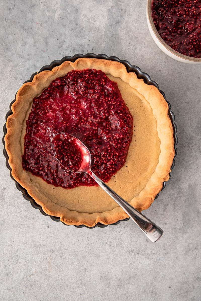 Overhead shot of the baked tart shell being filled with raspberry jam