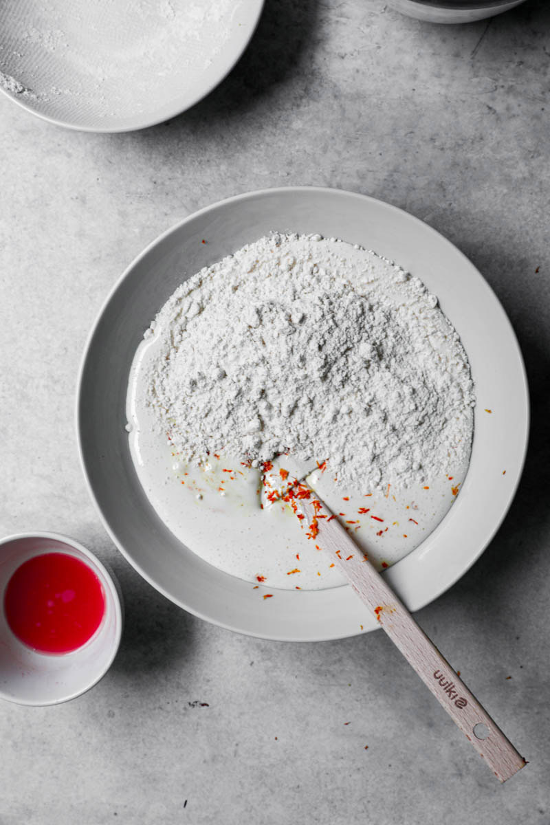 Blanched eggs and sugar plus blood orange zest in a white bowl with the flour and a wooden spatula.