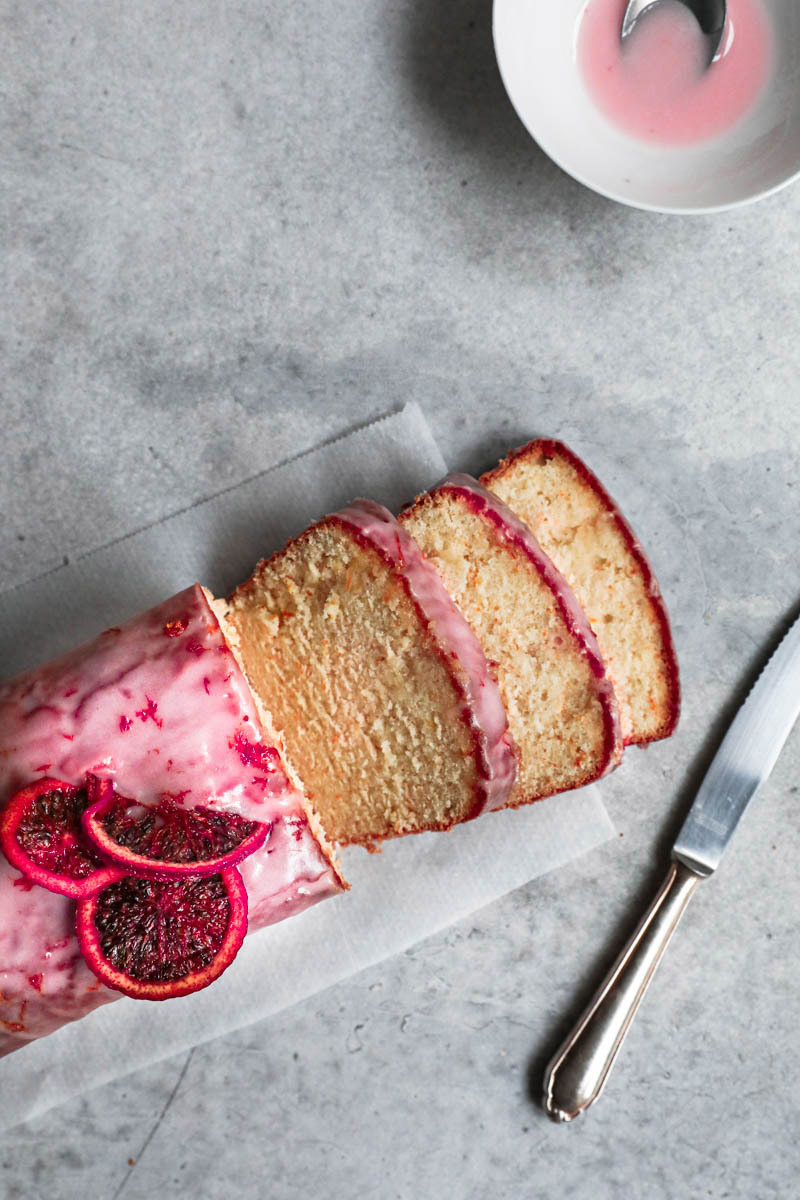 Sliced blood orange cake seen from above with a knife on the side and a bowl with glaze on the top right corner.