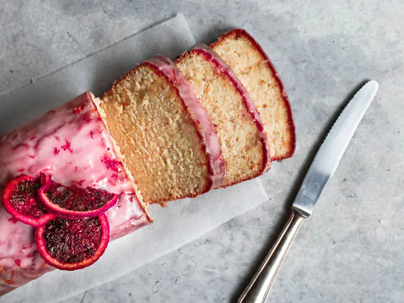 Sliced blood orange cake seen from above with a knife on the side and a bowl with glaze on the top right corner.