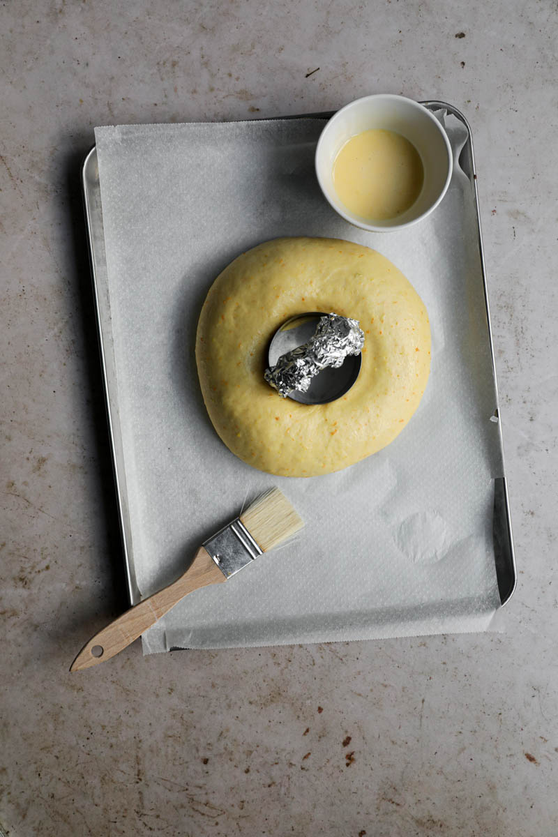 Easter bread with a whole in the middle and small cutter placed inside on a baking tray lined with parchment paper with a brush on the side and a small bowl with egg wash on the top right-hand side of the tray.