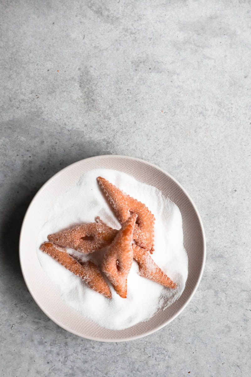 5 bugnes on a plate filled with granulated sugar