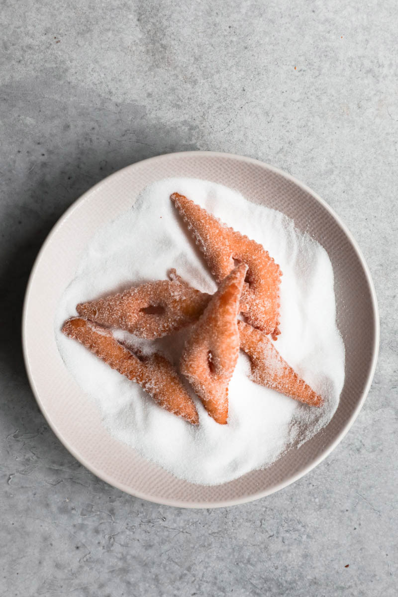 5 bugnes on a plate filled with granulated sugar