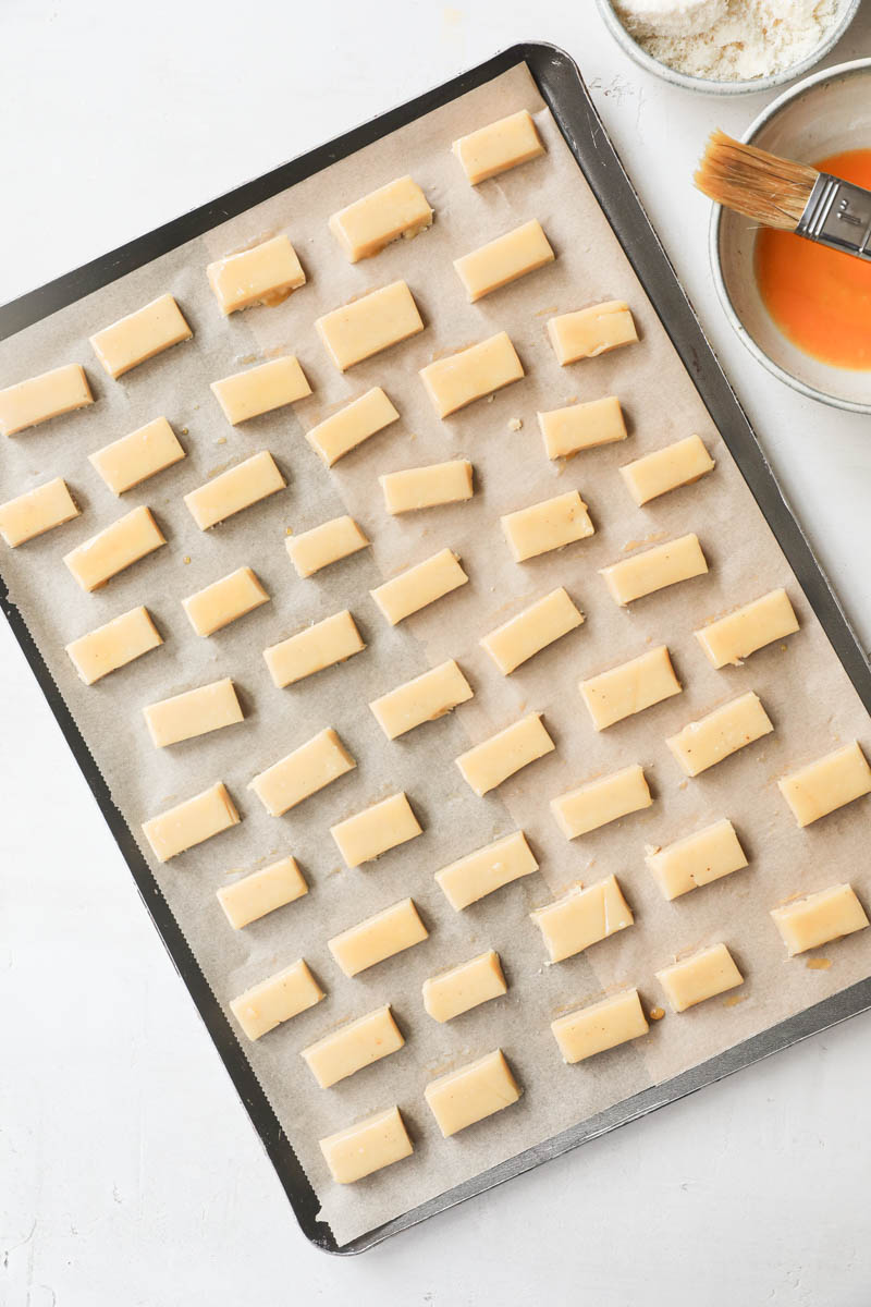 The parmesan cheese cookies placed on a baking tray lined with parchment paper brushed with egg wash with a bowl filled with egg wash and a brush on the side.