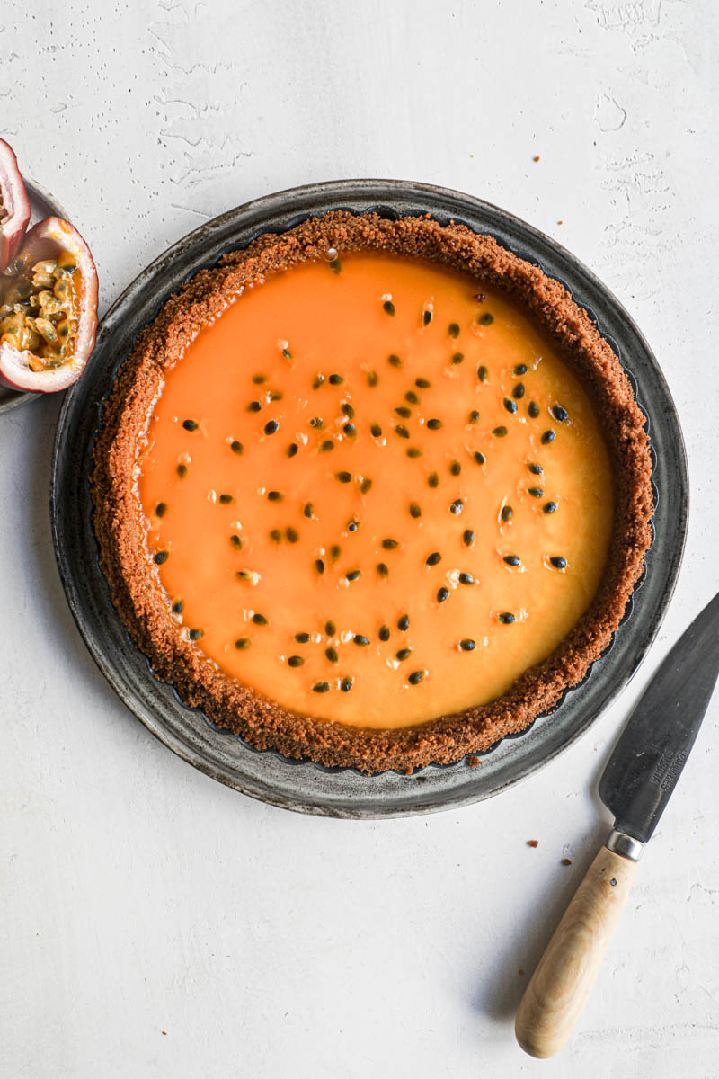 The passion fruit cheesecake tart on a grey plate with a knife on the side a passion fruit on the left top corner.