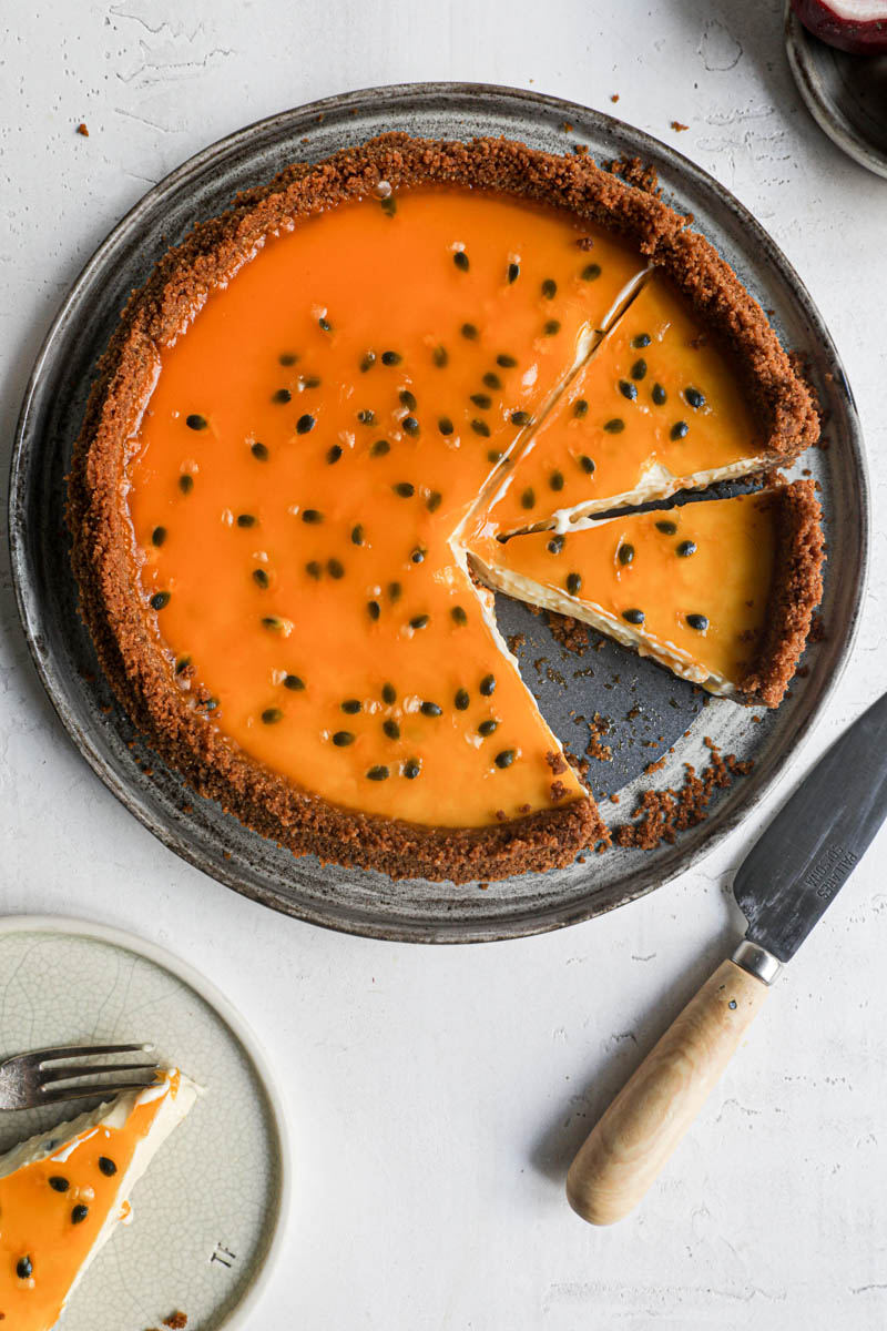 The sliced cheesecake tart as seen from the side on a grey plate with a passion fruit on the side of the frame and a small plate with a slice of cheesecake tart at the bottom of the frame.