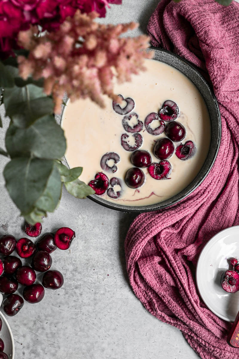 Overhead shot of the sliced cherries inside the sugared baking dish topped with the clafoutis batter with some flowers on the left upper corner