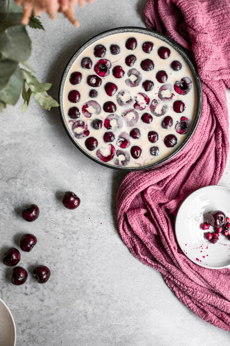 Overhead shot of the sliced cherries inside the sugared baking dish topped with the clafoutis batter with some flowers on the left upper corner
