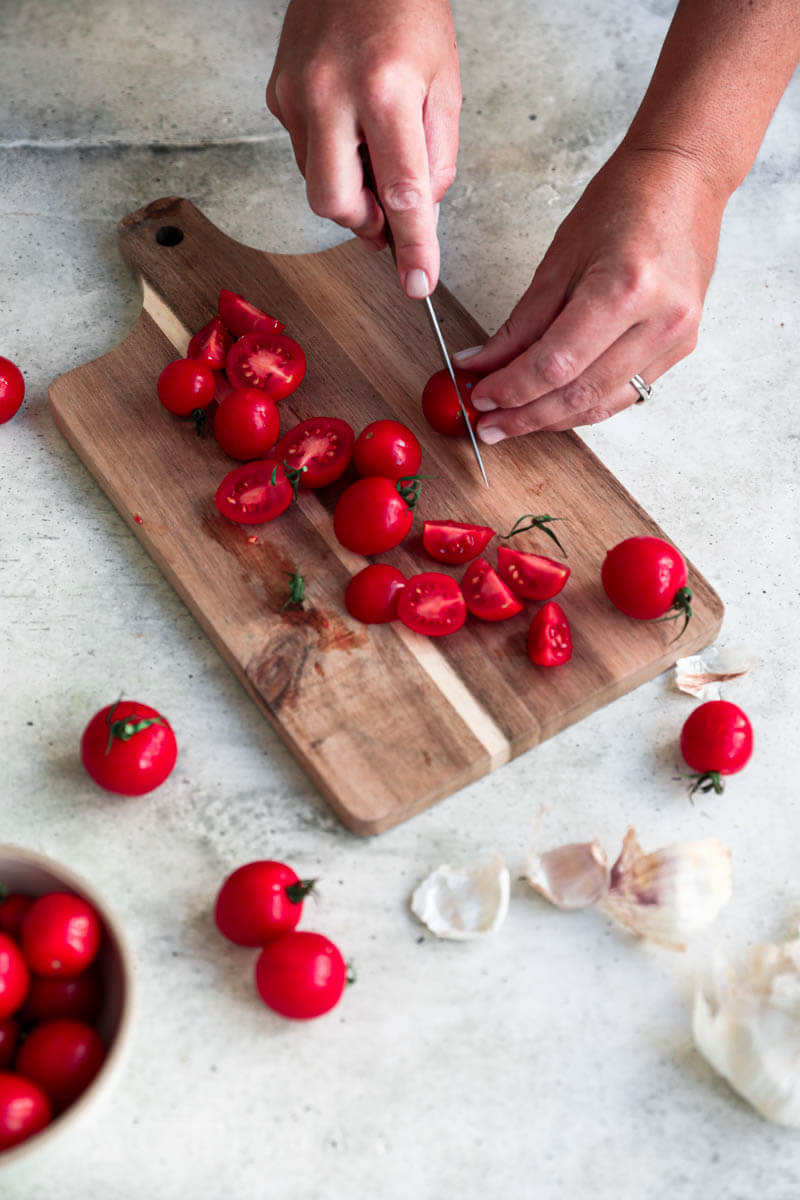 45° shot of 2 hands slicing cherry tomatoes on a wooden board