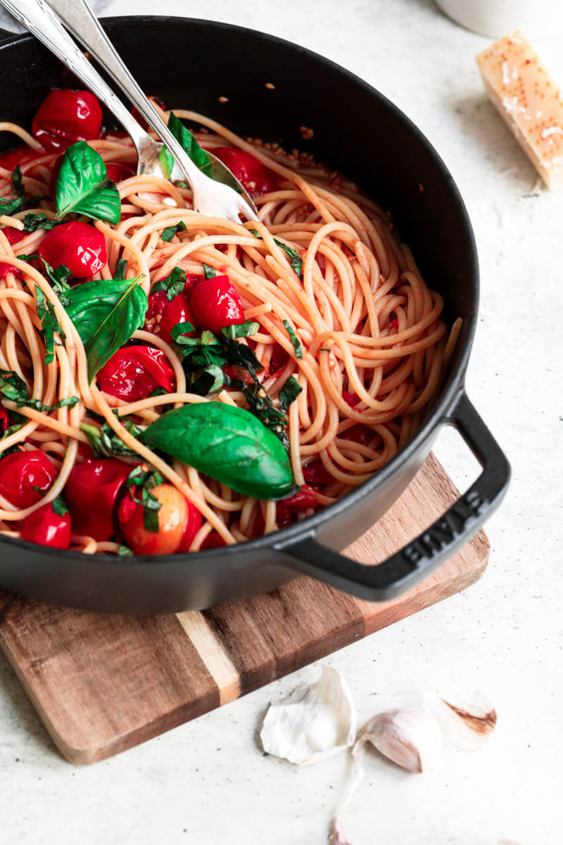 45° closeup shot of half a black pan filled with pasta and topped with cherry tomato sauce with a fork and a spoon in the corner