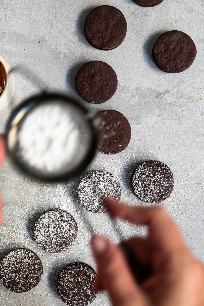 Overhead shot of a hand sprinkling powdered sugar onto the chocolate alfajores