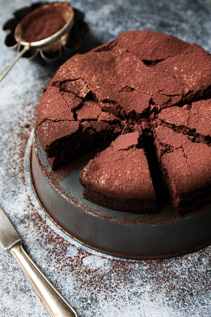 Sliced chocolate fudge on a grey tray with a knife on the side and a bowl with cacao powder in the back.