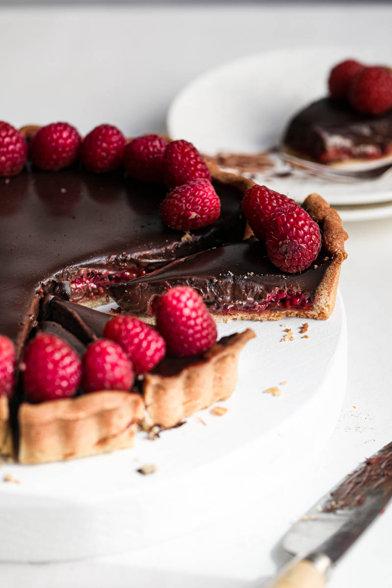 The sliced chocolate ganache raspberry tart on a white wooden board with a knife on the side.