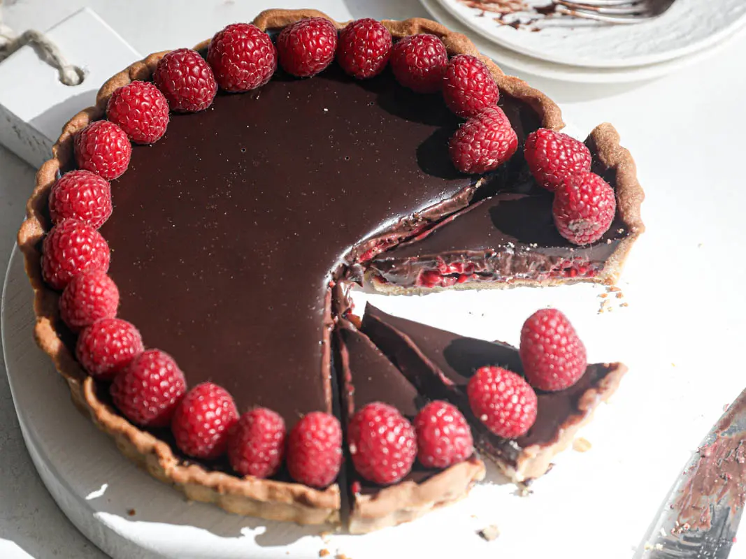 The sliced chocolate ganache raspberry tart on a white wooden board with a knife on the side.