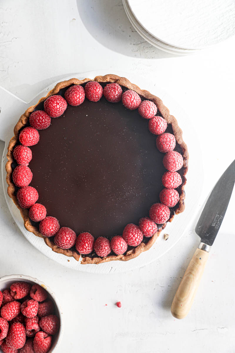 The chocolate ganache raspberry tart as seen from above with a knife int he side and a bowl of raspberries on the bottom left side of the frame.