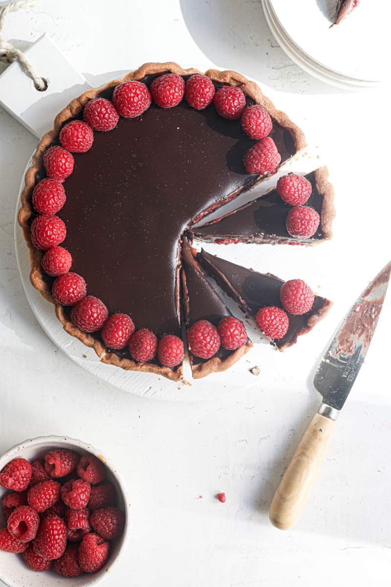 The sliced chocolate ganache raspberry tart as seen from above with a knife int he side and a bowl of raspberries on the bottom left side of the frame.