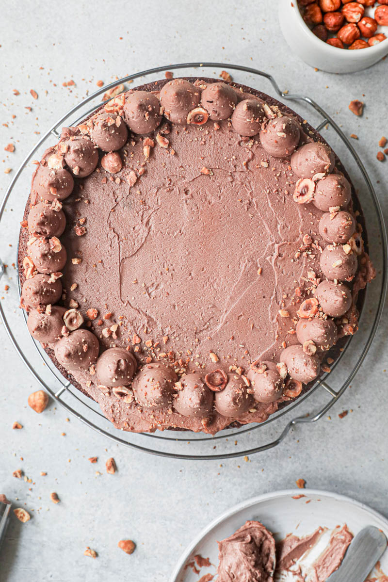 The chocolate hazelnut cake topped with whipped milk chocolate ganache on top of a wire rack with a plate on the bottom of the frame.