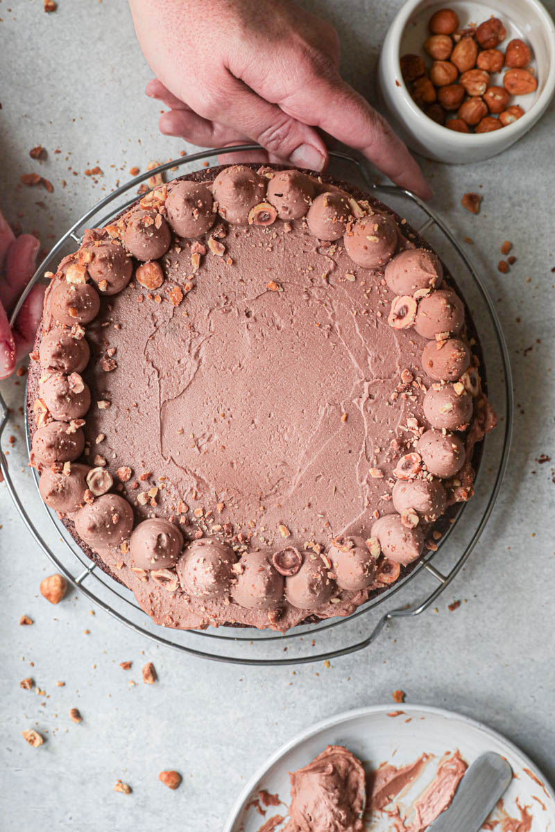 2 Hands holding the The chocolate hazelnut cake topped with whipped milk chocolate ganache on top of a wire rack.