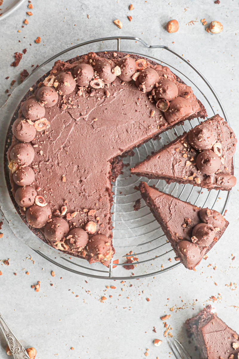 The sliced chocolate hazelnut cake topped with whipped milk chocolate ganache on top of a wire rack with a slice on the bottom right corner.