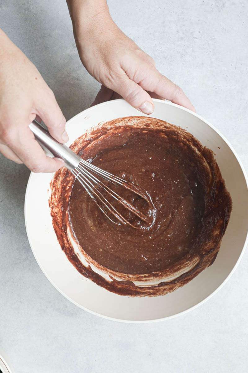 2 hands whisking in the chocolate/butter mixture into the chocolate hazelnut cake batter inside a beige bowl.