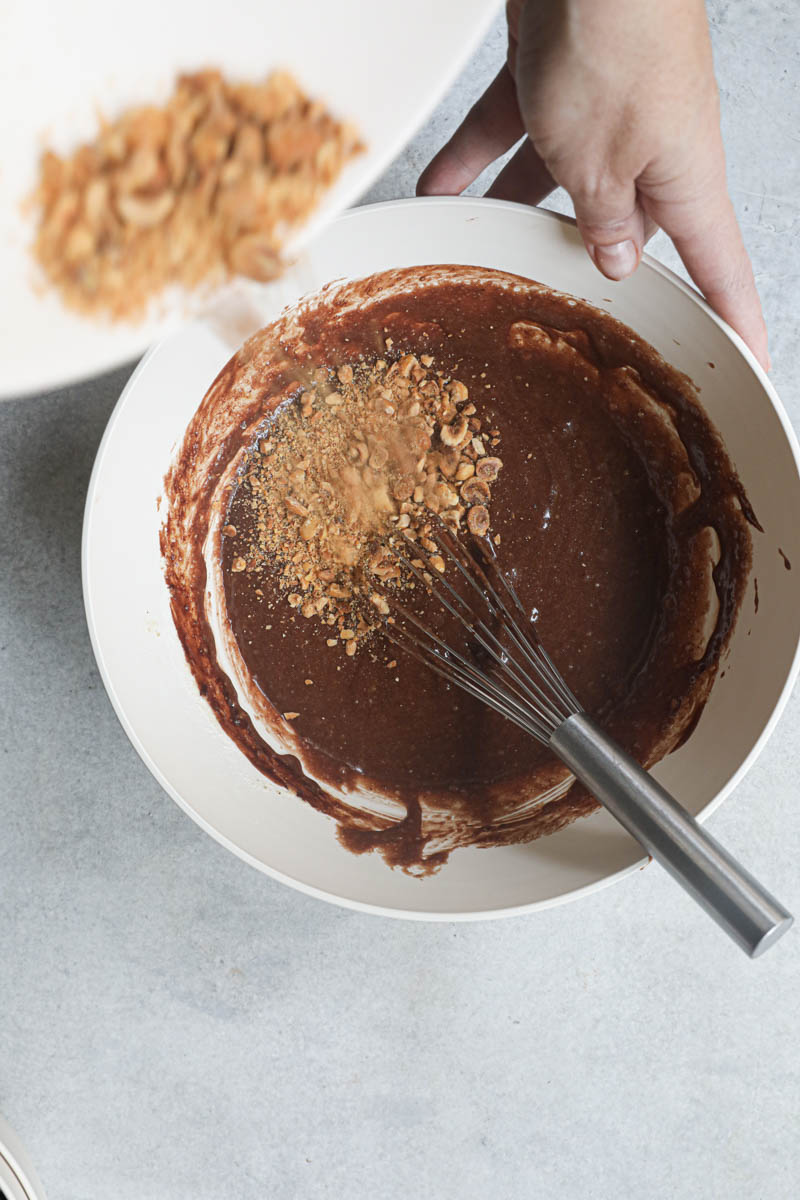 Chopped hazelnuts being added into the chocolate hazelnut cake batter inside a beige bowl.