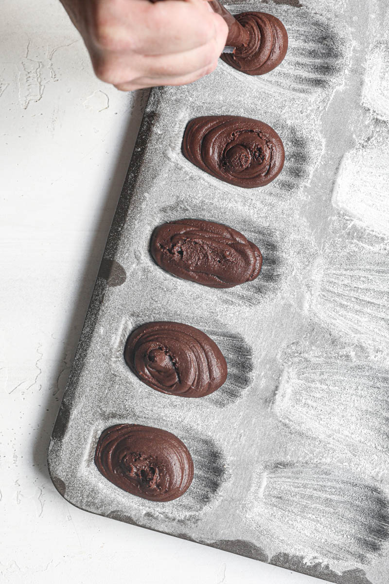 One hand ppiping the chocolate madeleine batter into the madeleine tray.