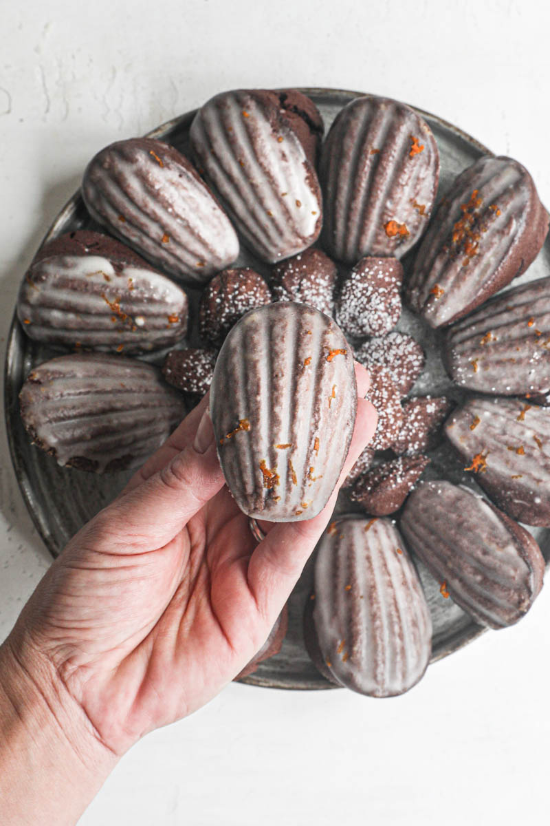 One hand holding a chocolate madeleine with the shell side up and other orange glazed madeleines underneath.