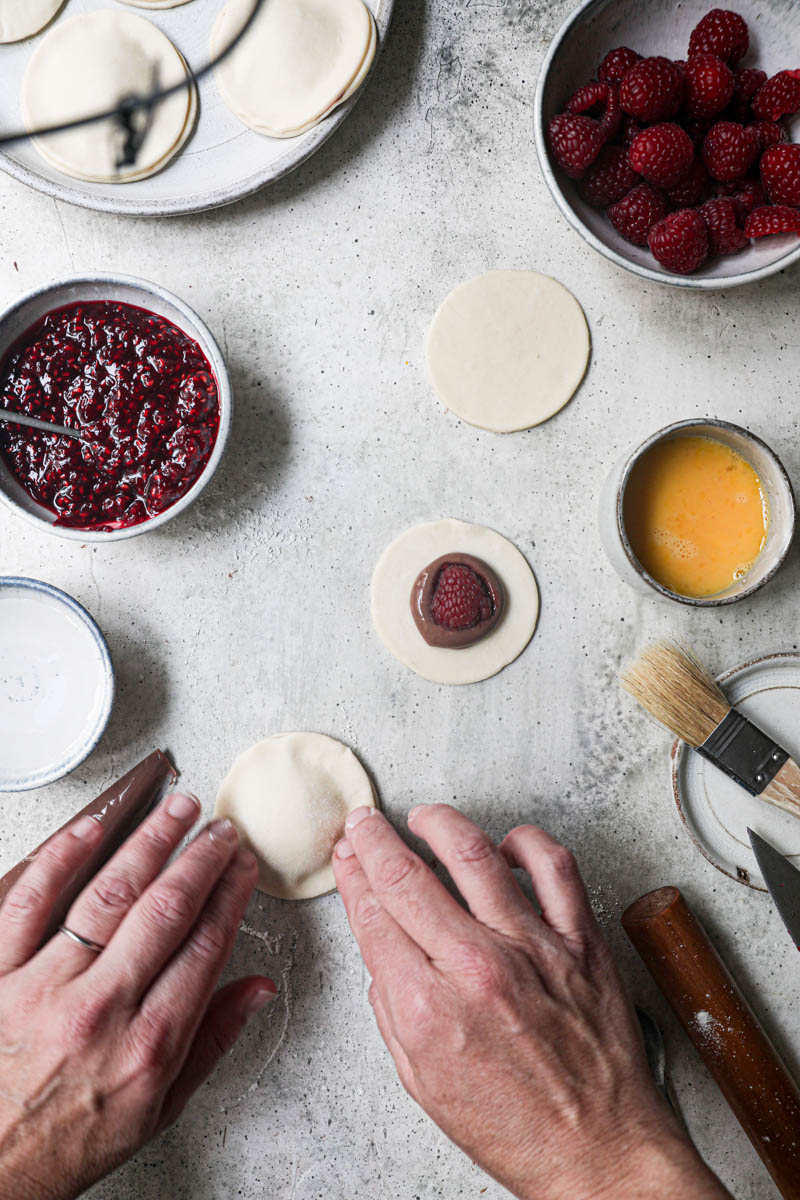 2 hands sealing a chocolate pudding hand pie sorrounded by fresh raspberries, already assembled hand pies on the top corner of the frame and a bowl with raspberyy jam on the side.