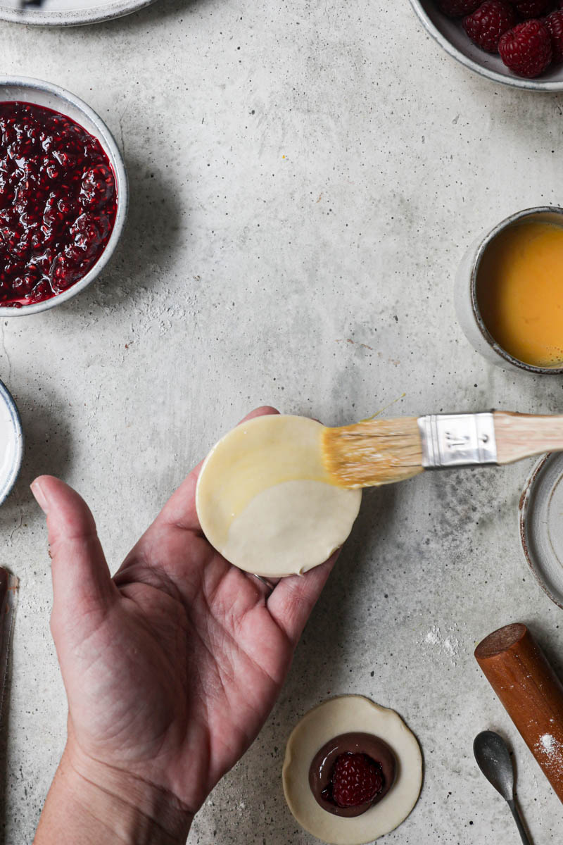 2 hands brushing a chocolate pudding hand pie with egg wash sorrounded by fresh raspberries, already assembled hand pies on the top corner of the frame.