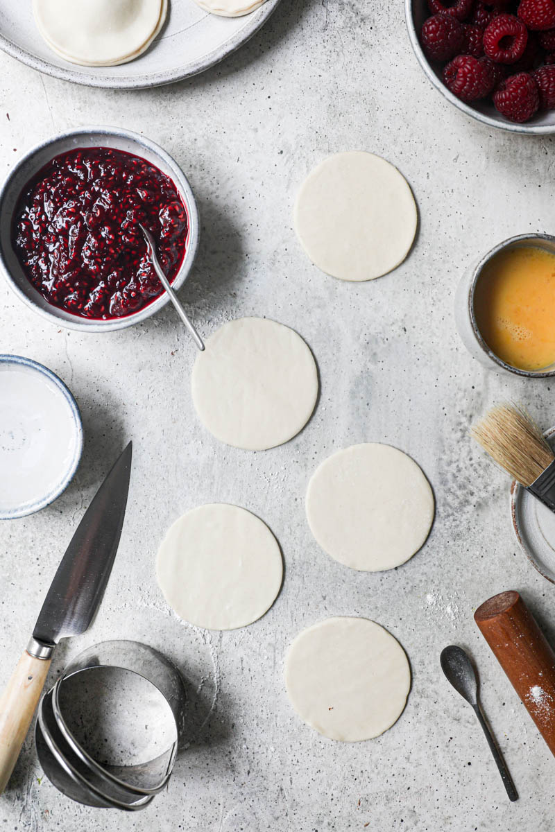 THe cut out pastry discs sorrounded by fresh raspberries, already assembled hand pies on the top corner of the frame and a bowl with raspberyy jam on the side.