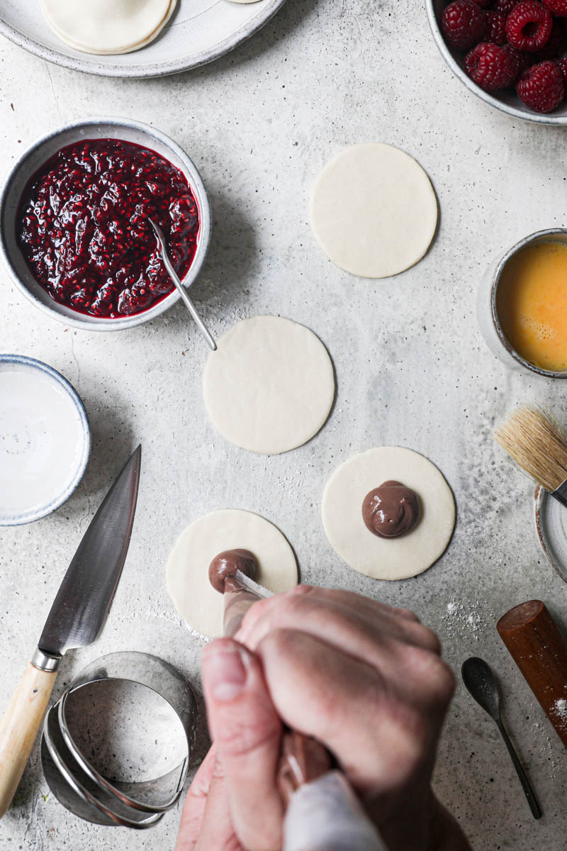 2 hands piping the chocolate pastry cream onto the cut out pastry discs sorrounded by fresh raspberries, already assembled hand pies on the top corner of the frame.