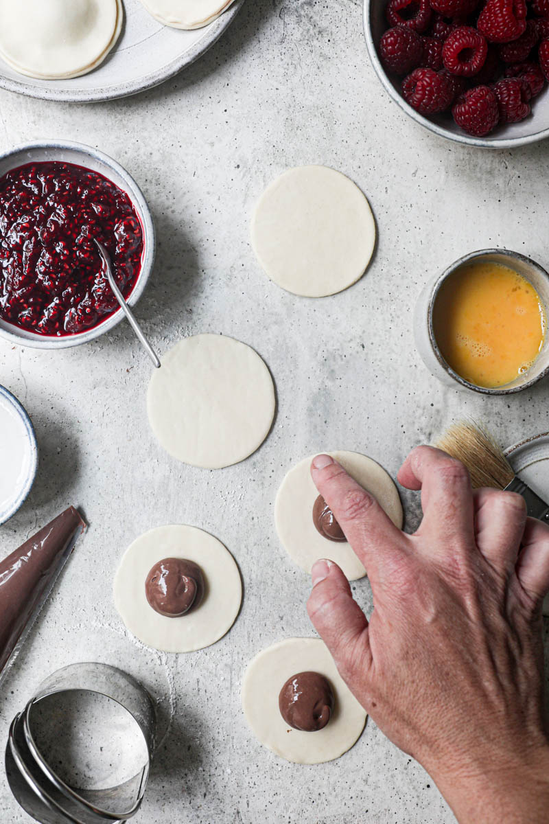 1 hand brushing water around the pastry discs filled with chocolate pastry sorrounded by fresh raspberries, already assembled hand pies on the top corner of the frame.