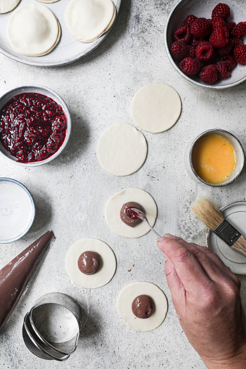 1 hand adding a dollop of raspberry jam onto the pastry discs filled with chocolate pastry sorrounded by fresh raspberries, already assembled hand pies on the top corner of the frame.