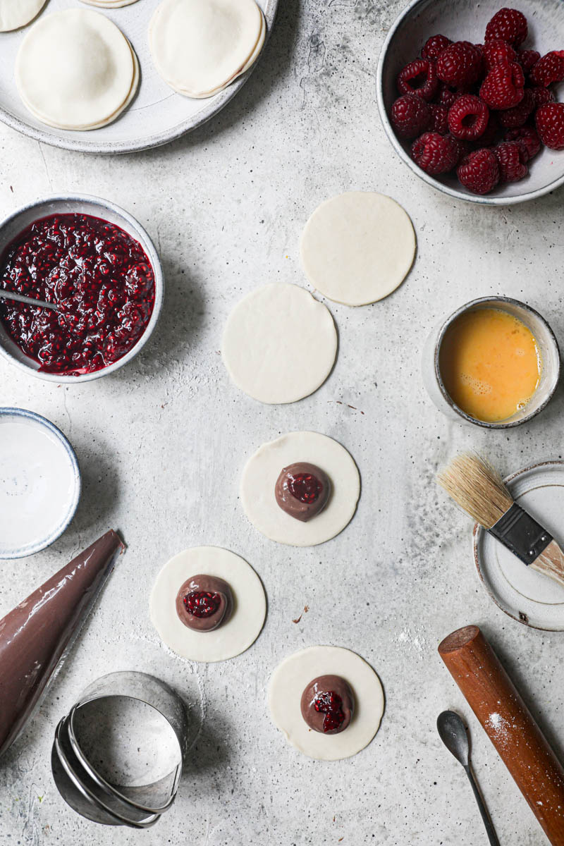 The pastry discs filled with chocolate pastry sorrounded by fresh raspberries, already assembled hand pies on the top corner of the frame and a bowl with raspberyy jam on the side.