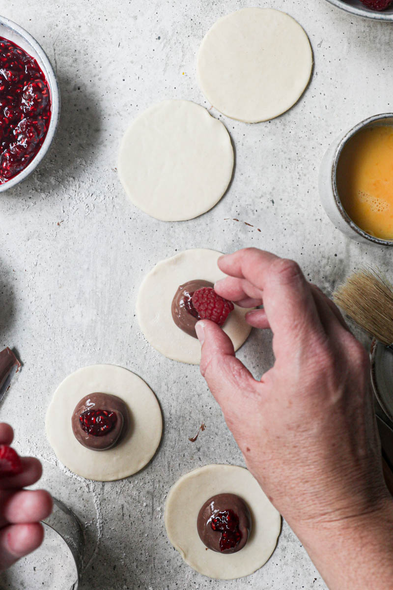 1 hand adding a fresh raspberry onto the pastry discs filled with chocolate pastry sorrounded by fresh raspberries, already assembled hand pies on the top corner of the frame.