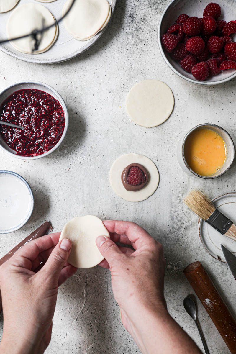2 hands placing a pastry disc onto the cut out pastry discs filled with chocolate pastry cream sorrounded by fresh raspberries, already assembled hand pies on the top corner of the frame.